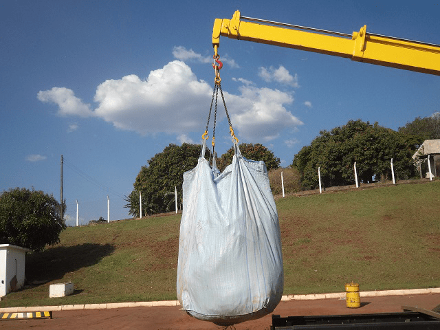 Lança de guindaste munck içando um big bag claro preso por correntes e ganchos (suporte para big bag), em área externa com gramado em declive e céu azul com nuvens.