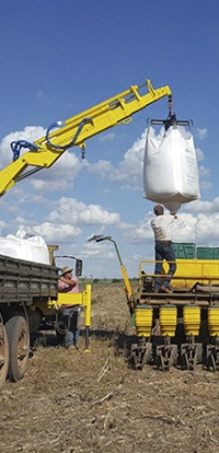 aplicação agrícola com munck Rodomunk suspendendo um big bag branco acima de uma plantadeira/amarela, enquanto um trabalhador em cima da máquina acompanha o abastecimento; cenário de lavoura com céu aberto e nuvens.