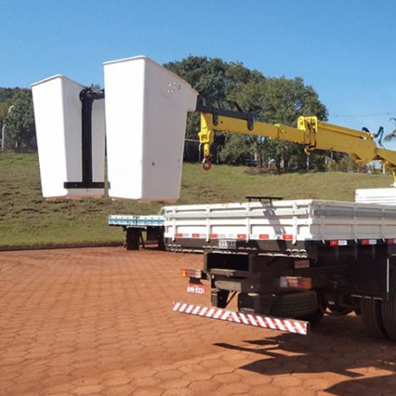 cesto de inspeção branco sendo içado pelo guindaste ao lado de um caminhão com carroceria, com estabilizadores (sapatas) abertos e operação em área externa pavimentada, indicado para manutenção/instalação em altura.