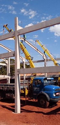 guindaste (munck) Rodomunk levantando um artefato de concreto de grande porte (peças tipo cochos/bebedouros ou pré-moldados) por cintas, com várias unidades similares empilhadas na carroceria; trabalhadores ao lado auxiliam no posicionamento em pátio de terra vermelha.