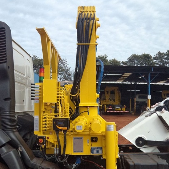 vista do conjunto de munck Rodomunk instalado no chassi do caminhão, com coluna amarela, feixe de mangueiras hidráulicas, painel/módulo de comando e componentes elétricos; ao lado, parte da cabine e do pátio/galpão ao fundo.