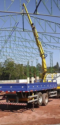 caminhão plataforma azul com guindaste Rodomunk trabalhando sob uma estrutura metálica de galpão (treliças verdes), com a lança elevada para auxiliar na montagem; estabilizadores apoiados no solo vermelho do canteiro