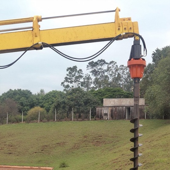lança do guindaste com kit de perfuratriz, mostrando o motor/acionamento (laranja) e a broca helicoidal apontada para o solo, pronta para perfurar em terreno rural com árvores e construção ao fundo.