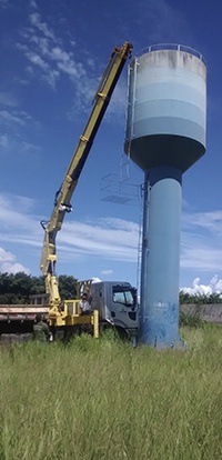 caminhão com guindaste Rodomunk estendido para realizar serviço em altura em uma torre de caixa d’água azul; a lança alcança próximo ao topo do reservatório, em área gramada sob céu claro.