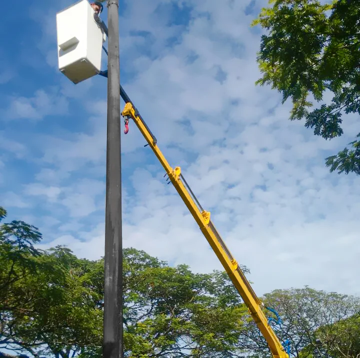 guindaste veicular (Munck) Rodomunk com lança estendida até um poste alto, utilizando cesto aéreo para elevar o técnico até a altura de trabalho; gancho vermelho visível na lança e céu parcialmente nublado com árvores preenchendo a parte inferior da imagem.