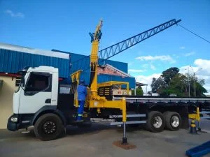 guindaste (munck) Rodomunk GRM 25000, para até 6,2 toneladas 4H em caminhão plataforma, com estabilizadores abertos, içando treliça/viga metálica longa (azul) para montagem de cobertura; operador em uniforme azul acompanha a manobra ao lado de galpão com fachada azul.