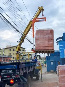 guindaste veicular (Munck) Rodomunk içando um pallet grande de matéria-prima (blocos/tijolos cerâmicos envoltos em filme) usando acessório tipo garfo/levantador de carga laranja com indicação de “2000 kg”; operação em rua com muitos cabos aéreos e trabalhadores acompanhando o descarregamento na carroceria do caminhão.