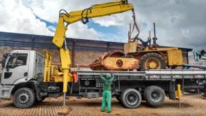 munck Rodomunk amarelo carregando máquina/equipamento pesado sobre caminhão prancha, preso por cintas; trabalhador de uniforme verde orienta a manobra ao lado da carga, com estabilizadores apoiados no chão e galpão industrial ao fundo.