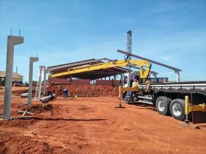 guindaste veicular Rodomunk com lança longa operando em obra de solo vermelho, posicionando/transportando perfis e elementos da estrutura enquanto equipe trabalha no canteiro; ao fundo, estrutura de cobertura em montagem e torre metálica vertical, com caminhão estabilizado por sapatas.