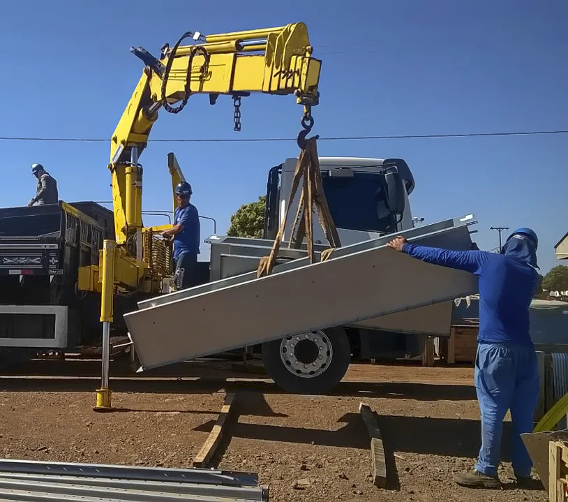 guindaste veicular Rodomunk içando uma peça metálica longa (painel/viga leve) por cintas, com trabalhador em azul segurando a extremidade para evitar balanço; operador e equipe na carroceria do caminhão acompanhando o descarregamento sob céu azul.