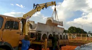 guindaste veicular (Munck) Rodomunk levantando cocho/bebedouro de concreto com formato curvo (peça volumosa) por cintas, sobre carroceria carregada com várias unidades; trabalhadores ao lado organizam o empilhamento em canteiro com terra vermelha e cerca ao fundo.