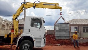 munck Rodomunk montado em caminhão branco içando um pallet de pavers/blocos intertravados embalado e cintado, com gancho e cintas em “V”; obra residencial ao fundo e trabalhador com capacete observando a descarga em terreno de terra vermelha.