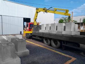 guindaste veicular Rodomunk amarelo operando em pátio industrial, com caminhão carregado de grandes blocos/peças de concreto na carroceria; galpão metálico ao fundo e várias unidades empilhadas, indicando movimentação e logística de artefatos de concreto.