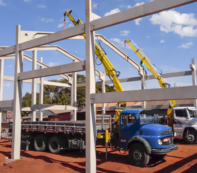 dois caminhões com munck Rodomunk trabalhando simultaneamente na montagem de estrutura pré-moldada (pórticos e vigas de concreto), com braços amarelos erguidos dentro do esqueleto da construção; cenário de obra com solo vermelho e céu com nuvens.