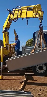 Cena de obra com guindaste veicular amarelo içando caçamba/caixa metálica cinza com cintas, com operador guiando a movimentação ao lado do caminhão.