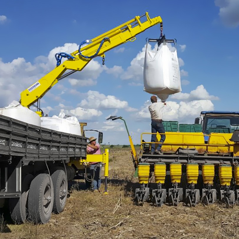 guindaste veicular amarelo em operação no campo, içando big bag de insumos/sementes e posicionando sobre uma plantadeira/amarela; dois trabalhadores acompanham a manobra em lavoura sob céu com nuvens.