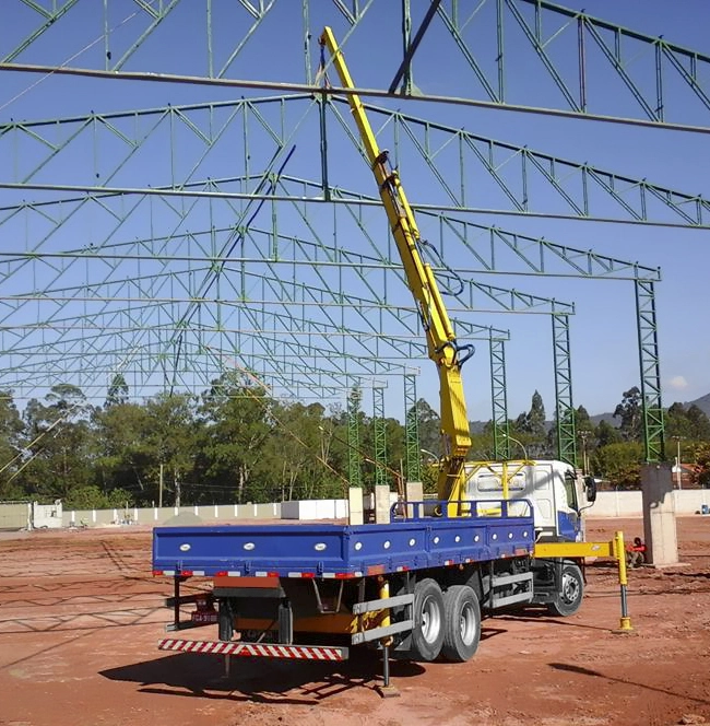 Caminhão com guindaste veicular (munck) amarelo operando em obra, com sapatas estabilizadoras abertas, içando/posicionando peças na montagem de estrutura metálica de galpão (treliças verdes) sob céu azul.