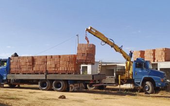 Guindaste veicular levantando carga de tijolos em obra, operação de içamento que exige comunicação do sinaleiro para orientar o operador.