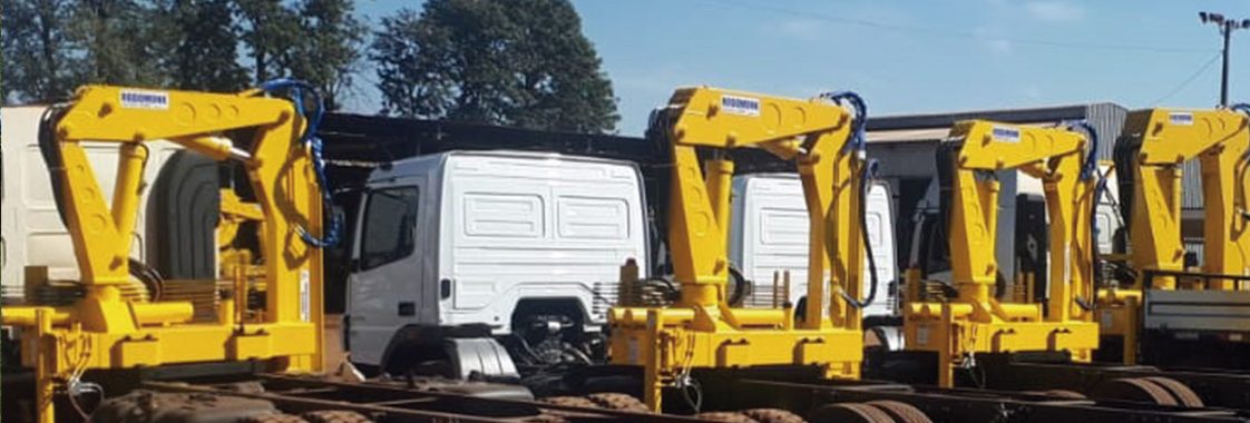 Guindastes veiculares articulados instalados em caminhões no pátio industrial, equipamentos que podem operar com controle remoto ou alavancas manuais.