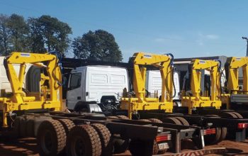 Guindastes veiculares articulados instalados em caminhões no pátio industrial, equipamentos que podem operar com controle remoto ou alavancas manuais.
