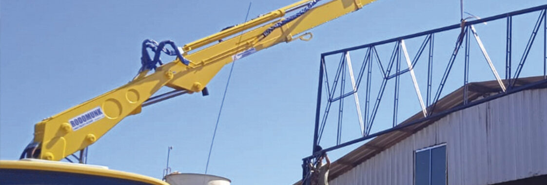 Guindaste veicular com lança estendida instalando estrutura metálica no topo de um galpão, demonstrando o alcance horizontal do equipamento sem reposicionar o caminhão.