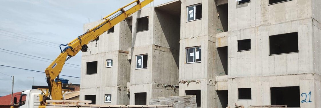 Guindaste veicular realizando o carregamento de peças pré-moldadas em um canteiro de obras, com patolas estabilizadas e operação próxima a um edifício em construção, ilustrando a etapa de içamento e posicionamento seguro das estruturas.