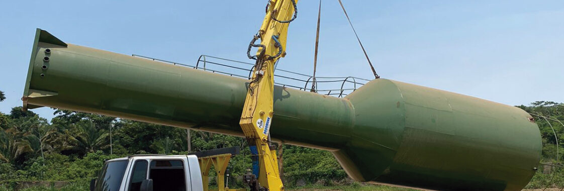 Guindaste veicular acoplado a caminhão realizando o içamento de uma caixa d’água de grande porte para posicionamento preciso na obra, demonstrando o uso do equipamento para elevação segura e controlada do reservatório.