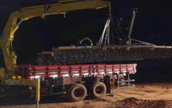 Guindaste veicular realizando içamento noturno de estrutura metálica sobre caminhão, com uso de iluminação artificial para garantir visibilidade e segurança na operação.