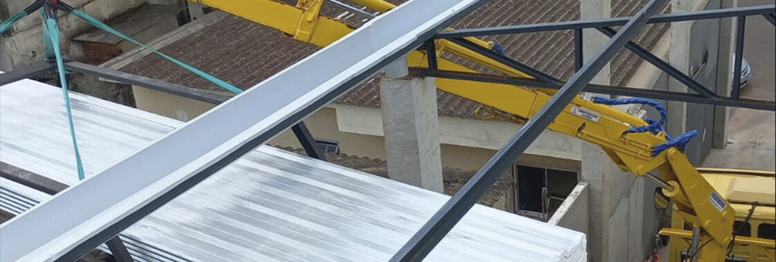 Guindaste veicular operando em espaço urbano reduzido, içando chapas metálicas sobre estrutura próxima a telhados e edificações, demonstrando a necessidade de precisão, controle de alcance e atenção a obstáculos ao redor.