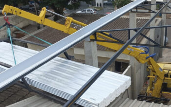 Guindaste veicular operando em espaço urbano reduzido, içando chapas metálicas sobre estrutura próxima a telhados e edificações, demonstrando a necessidade de precisão, controle de alcance e atenção a obstáculos ao redor.